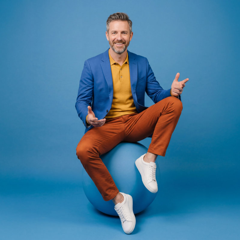 Mittelalter Mann mit Bart, gekleidet in orangefarbener Hose, gelbem Shirt und blauem Jacket, sitzt vor blauem Hintergrund auf einem blauen Würfel
